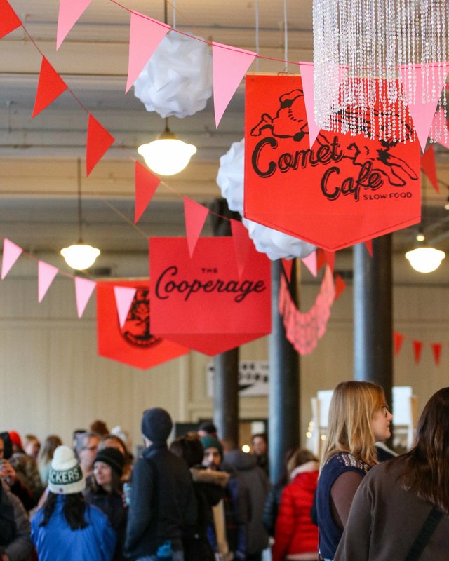 Shoppers crowd a bustling craft fair under pink and red pennant flags and hanging signs.