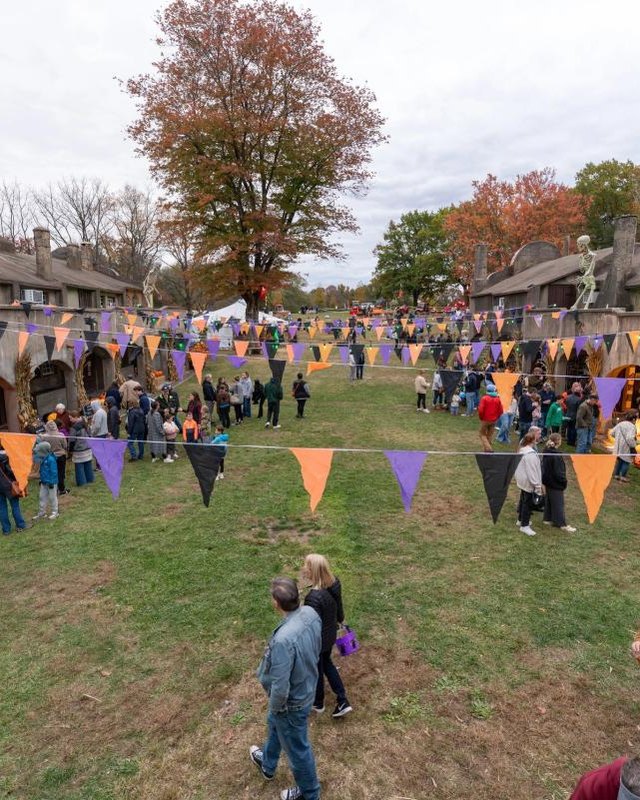 Crowd on a grass lawn at an autumn festival, with orange and purple bunting between gray stone buildings with arched doorways at the TileWorks.