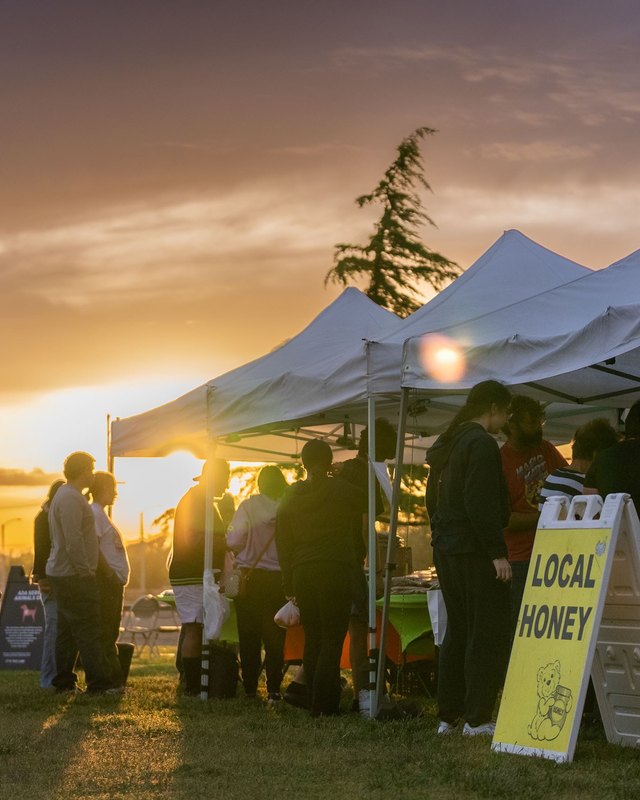 Evening market with white tents, shoppers silhouetted at sunset, and a Local Honey sign on the grass.