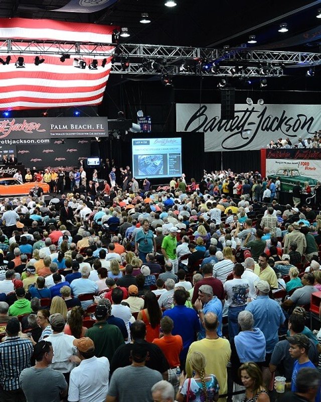 Large crowd packed into the Barrett-Jackson auction hall beneath an American flag and event signage.