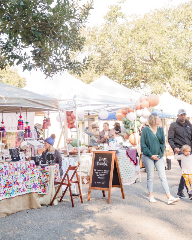 Market tents with shoppers walking past vendor booths under oak trees at an outdoor market.