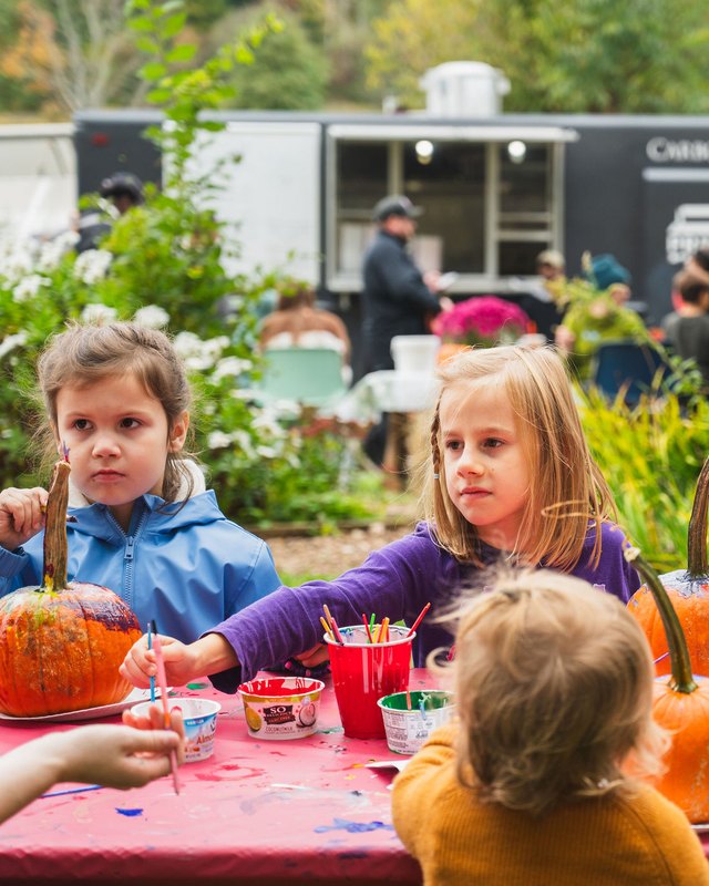 Children decorating pumpkins at a pink table, with a food truck and a person in a cap in the background at Auerfarm