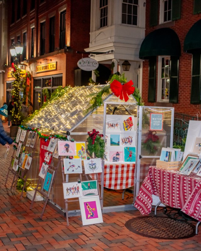 Holiday vendor booth with art prints under string lights on a Pratt Street brick sidewalk
