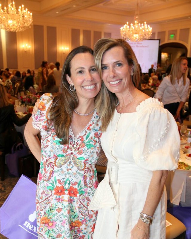 Two women posing together at a brunch: one in a floral print dress, one in a white dress, with chandeliers and a purple event tote bag nearby.