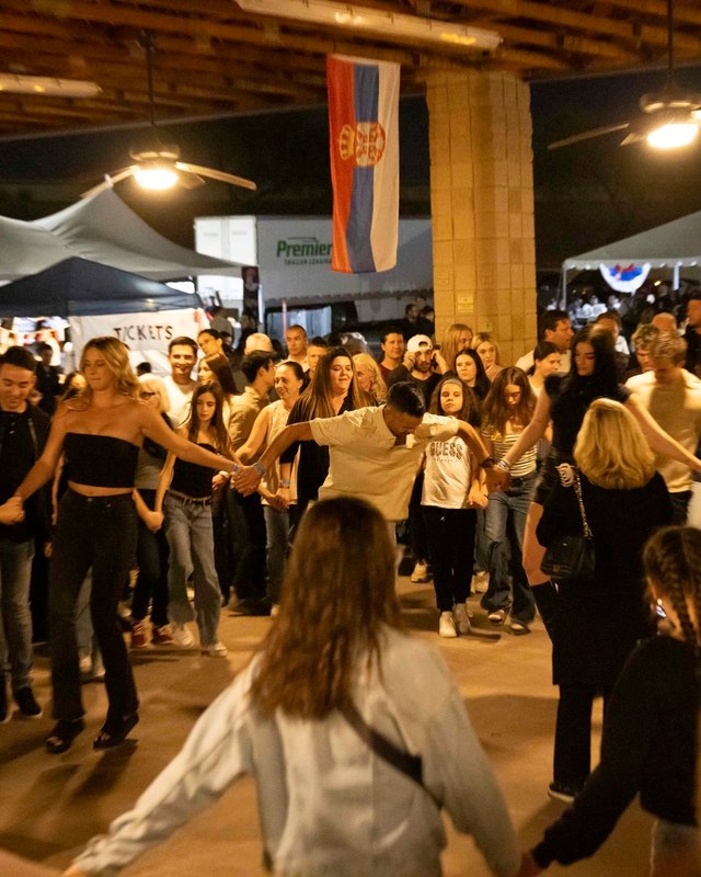 Crowd dancing at Serbfest Phoenix in front of tents and a Serbian flag at night.