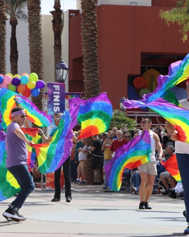 Dancers waving rainbow flags at the Cathedral City LGBT+ Days street festival, with balloons overhead.