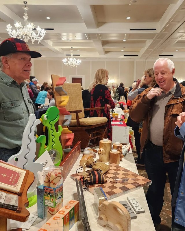 Visitors and vendors talking at a booth with crafts and goods inside a hotel ballroom at a holiday expo.