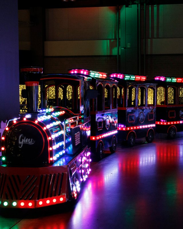 Illuminated trackless train with red and green LED lights on a glossy floor inside a large dark hall, with a glowing light display behind it.
