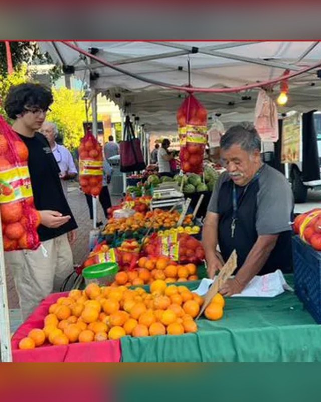 Shoppers and vendors at a produce stand with oranges and fruit under a white canopy at the Covina Farmers Market