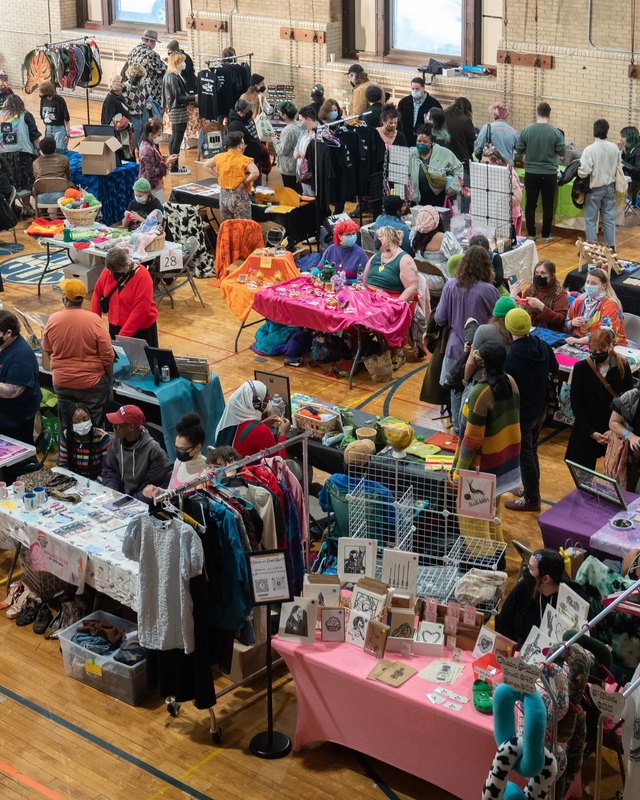 Shoppers browsing crafts and clothing tables at a crowded indoor market hall