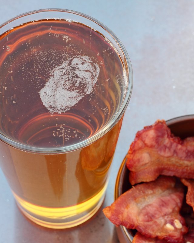 Glass of beer beside a bowl of cooked bacon on a light surface
