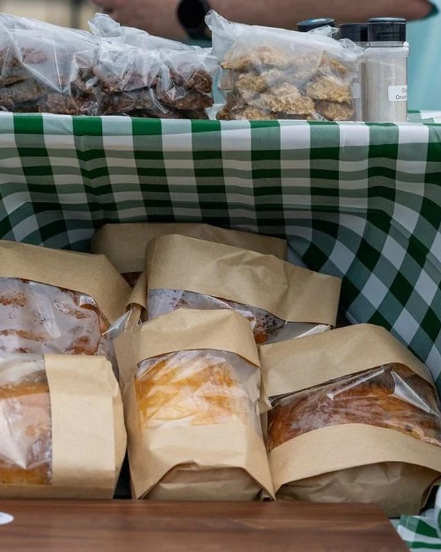 Bread in kraft paper bags and plastic tubs of green food on a green gingham tablecloth at a market table.