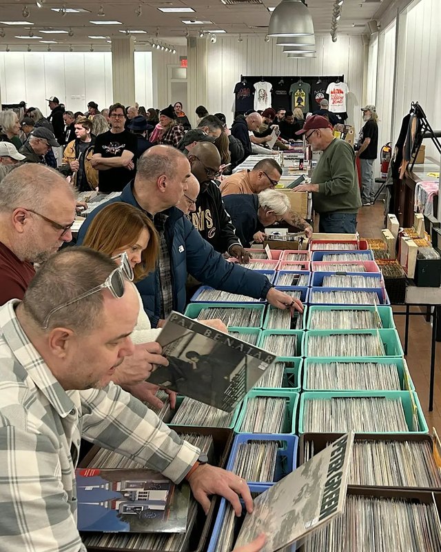 Shoppers flipping through record crates at tables of vinyl inside a bright indoor sale hall.