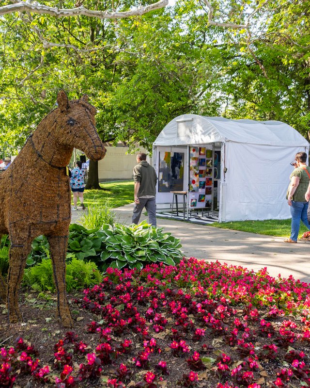 Vendor tents and shoppers along a park path beside a large rust-colored horse sculpture and red flower beds during JumpstART Weekend.