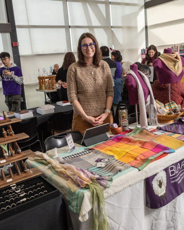 Makers Fair vendor table with wooden shelves of handmade goods and a woman standing behind it.