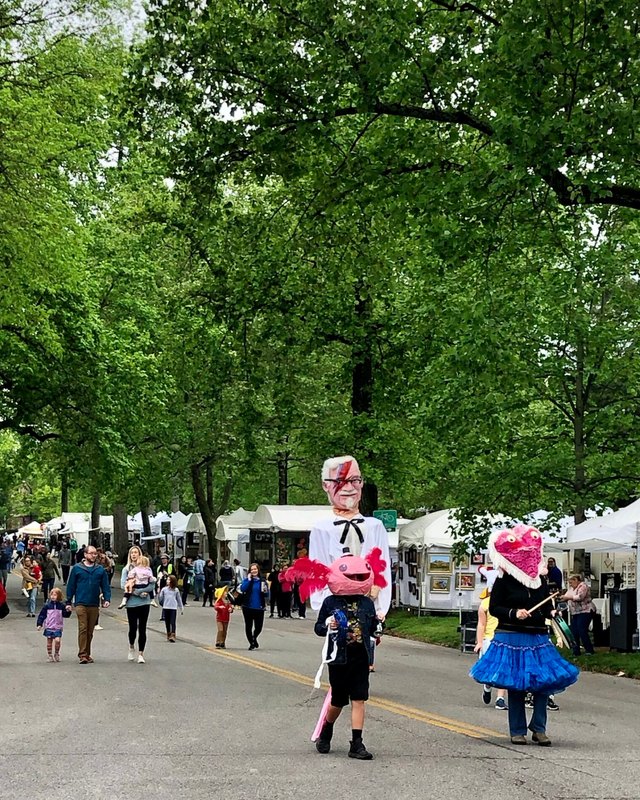 Festival path lined with white booth tents under trees, with costumed visitors walking among the crowd at the Cherokee Triangle Art Fair.