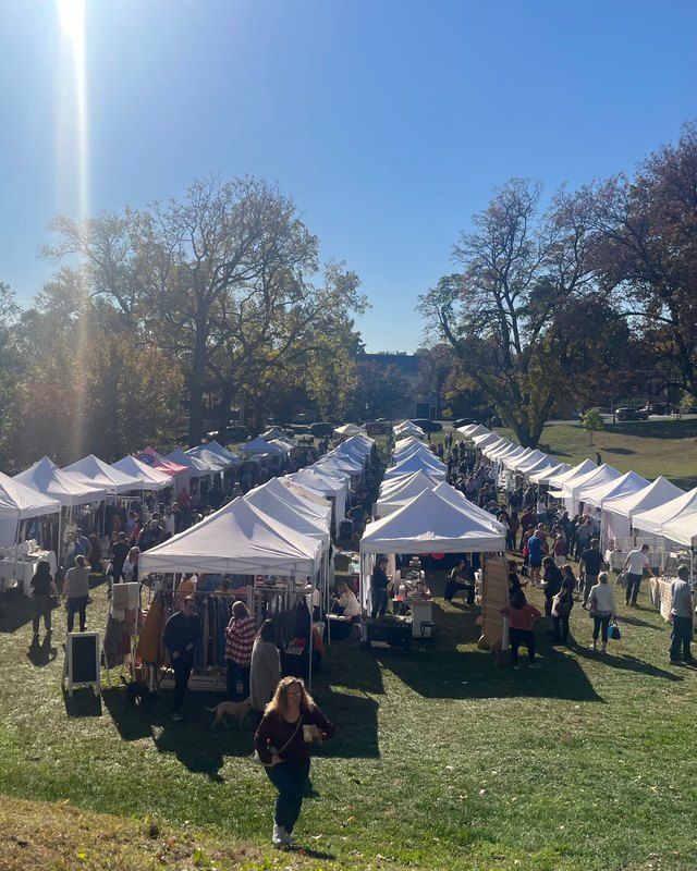 White tent booths and shoppers on a grassy lawn at an outdoor market under autumn trees.