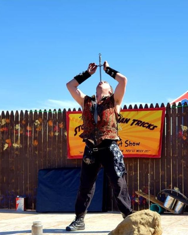 Performer with a sword act in front of a wooden fence and a Sword and Fun Tricks sign at the faire