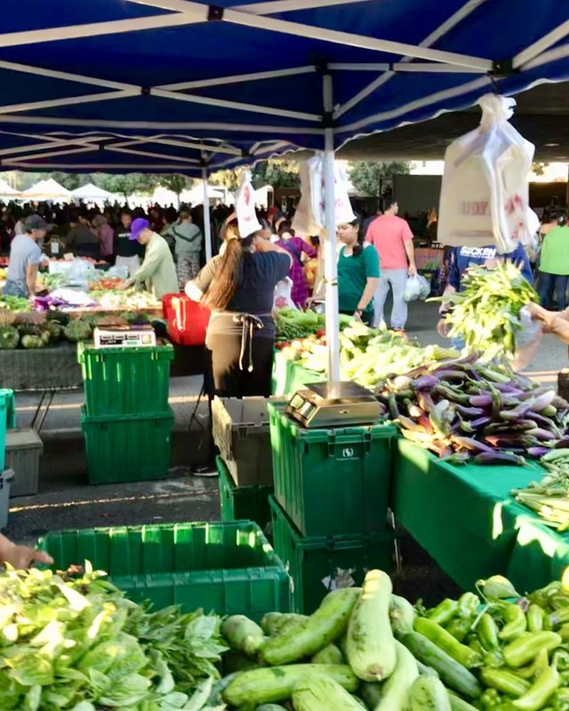 Shoppers and vendors at produce stalls under a blue canopy at the Golden Villa Farmers' Market