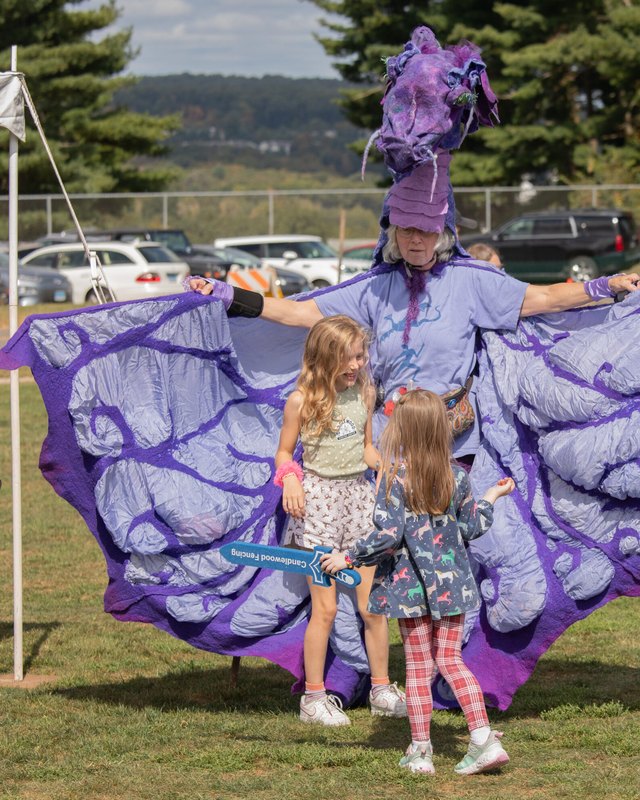 Costumed stilt performer in a large purple butterfly costume with two children outdoors at the festival