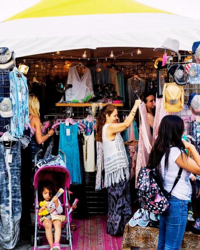 Shoppers browsing clothing and hats at a colorful festival vendor booth under a yellow canopy.