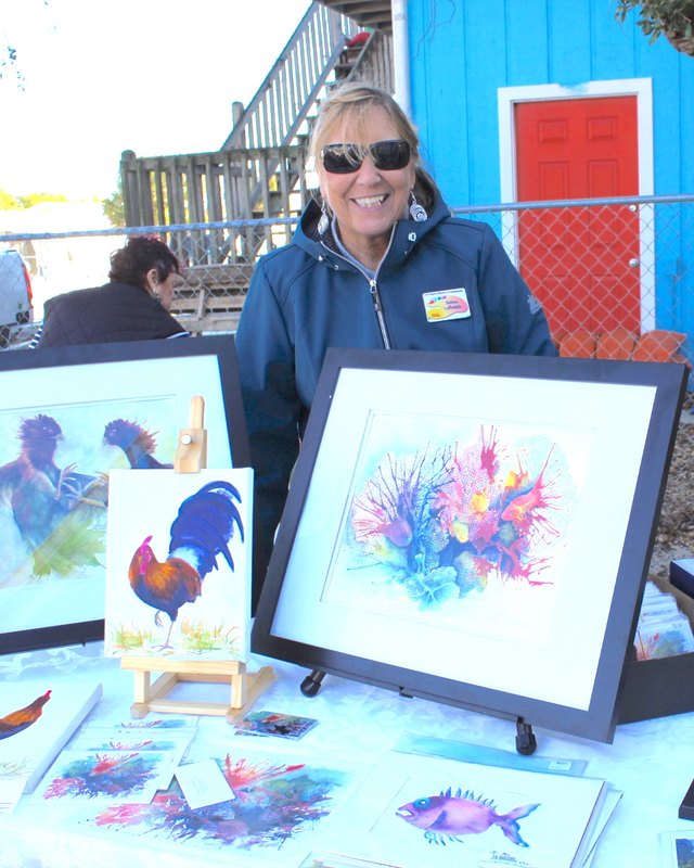 A smiling woman in a blue jacket and sunglasses stands at a booth with framed watercolor paintings and cards on easels, beside a blue building.