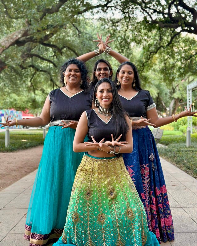 Four women in colorful lehengas and black dance-company T-shirts posing with arms raised under trees at Discovery Green in Houston.