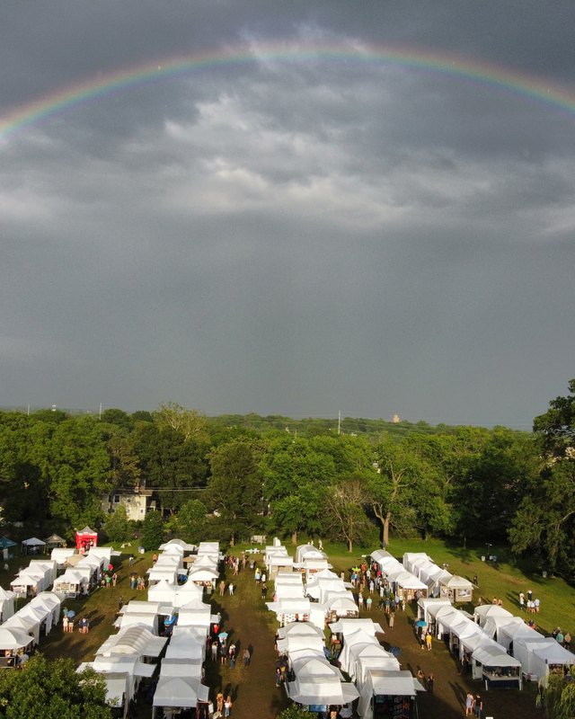 Aerial view of white vendor tents spread across a green lawn under a rainbow at the Webster Arts Fair