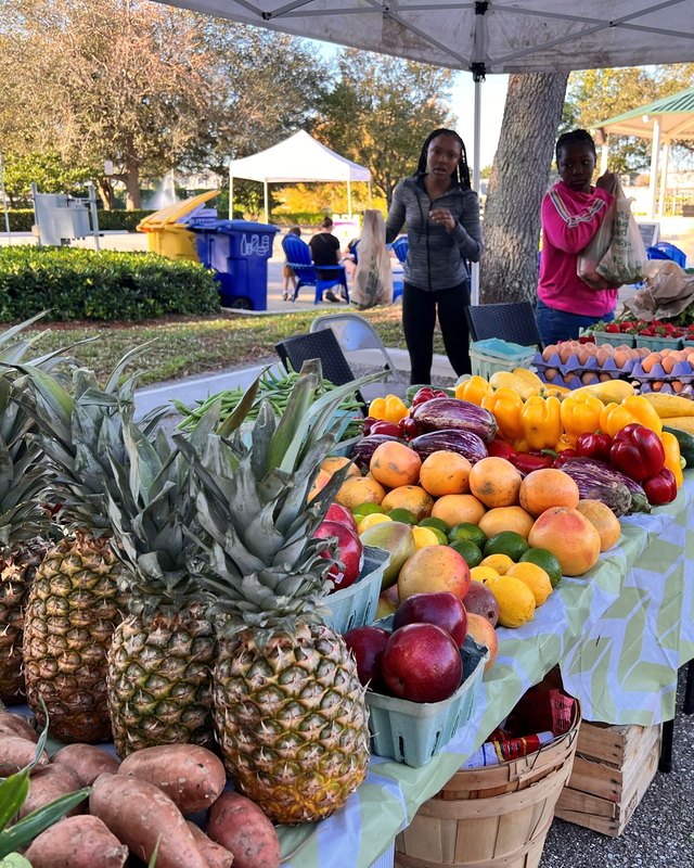 Shoppers at a market stall piled with pineapples, oranges and produce under a canopy.