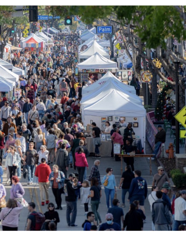 Crowds walking past white tent booths on a tree-lined street at an outdoor art fair
