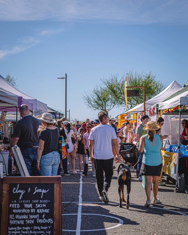 Shoppers walking between market booths at the Eastmark Farmers Market under a clear blue sky.
