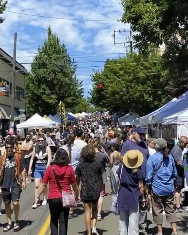 Dense crowd of fair visitors walking past white vendor tents on a tree-lined street