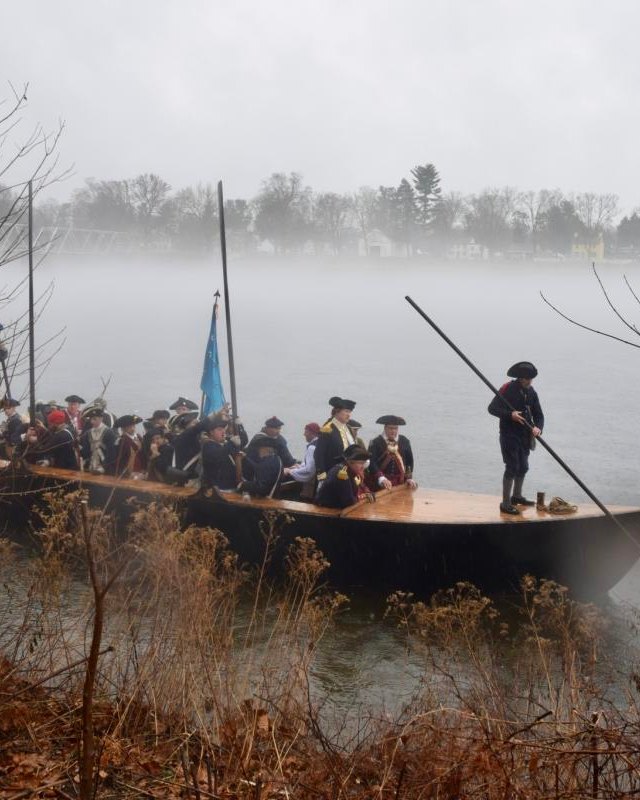 Reenactors in colonial dress on a flat-bottomed boat on a misty river, with bare trees along the bank.