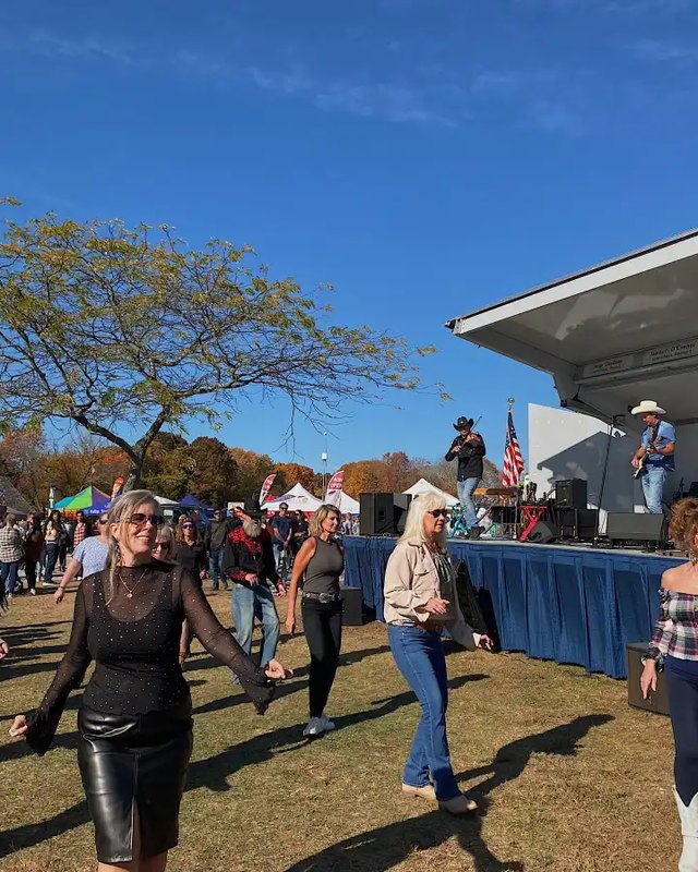 Women dancing on a sunny lawn in front of a stage with a blue skirt and musicians, with striped tents and a crowd behind them at the Islip Apple Festival.