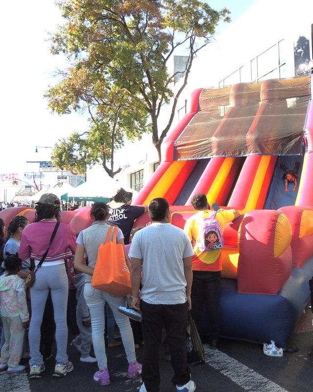 Street fair crowd on a closed avenue around an orange inflatable slide, with storefronts behind.