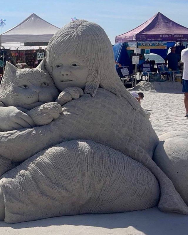 Sand sculpture of a child hugging a large cat, with vendor tents and visitors behind it on the beach.
