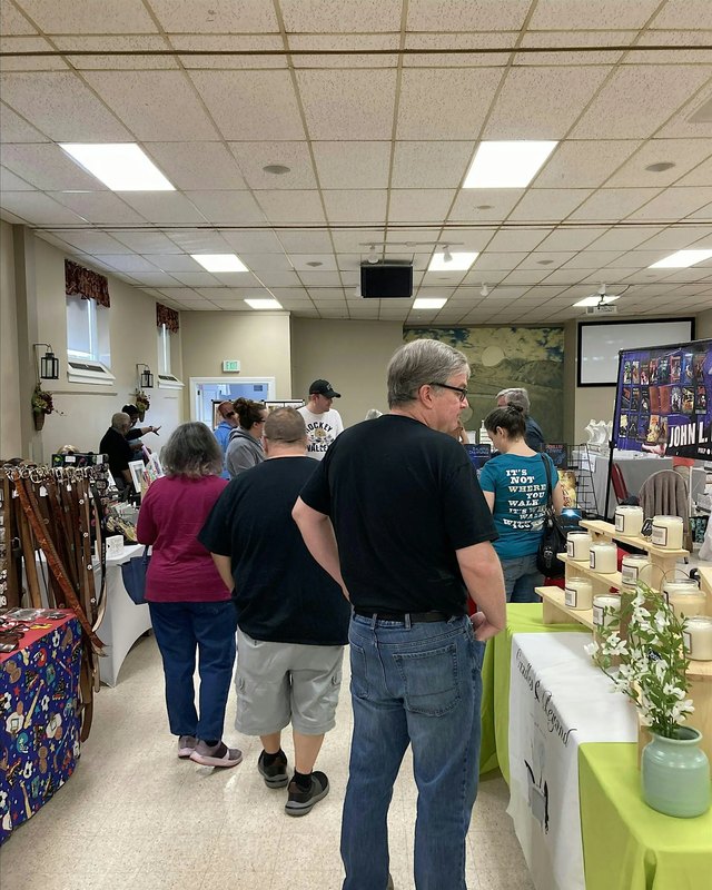 Shoppers browsing craft booths in a bright indoor market room with tables and displays.