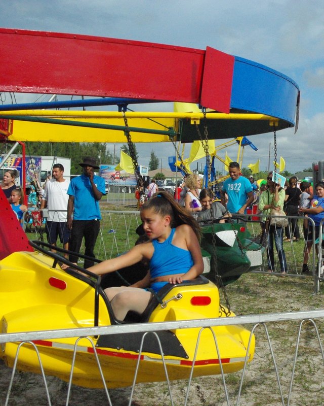 A girl in a blue top rides a yellow pedal attraction at the KidsFest festival, with crowds and yellow flags behind metal barriers.