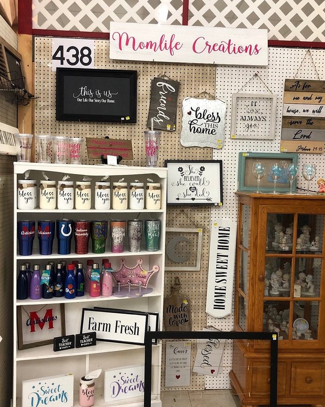 Momlife Creations booth at the flea market, with mugs, signs and home decor on pegboard shelves.