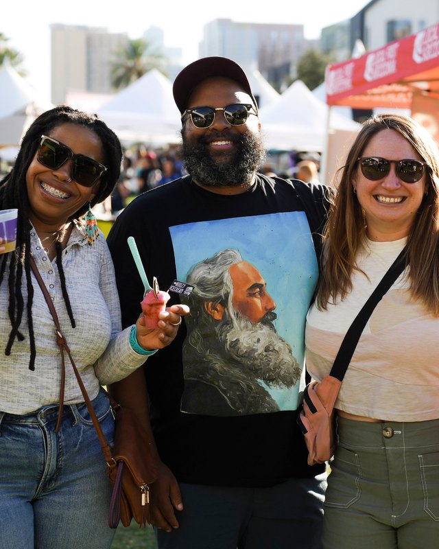 Three smiling festival visitors in sunglasses posing outdoors at a Local First Arizona event, with white tents and city buildings behind them.