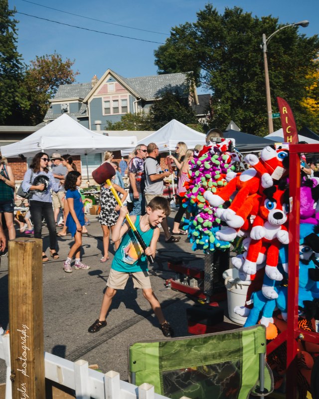 Crowd walking past street-fair tents and a prize booth stocked with plush toys during Bay View Bash