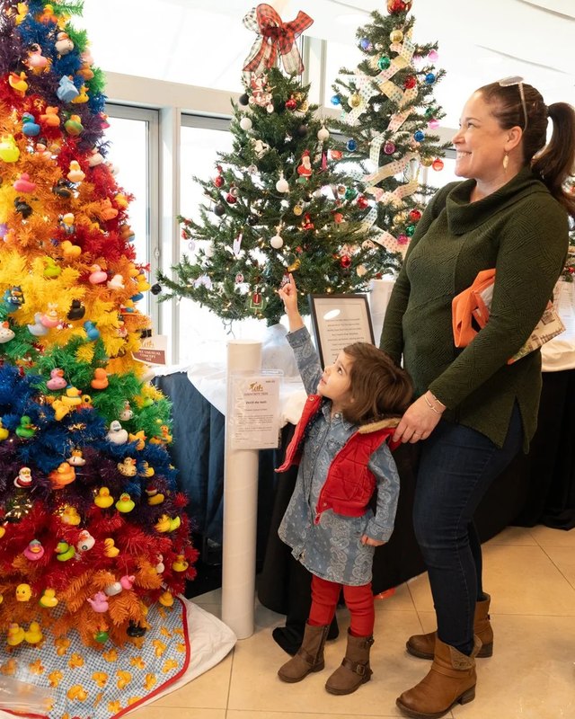 Visitors admiring decorated trees at the Festival of Trees, with a woman and a young child in front of a colorful tree.