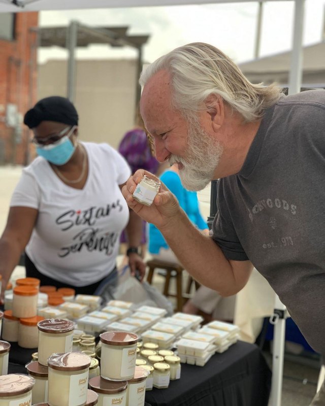 Vendor at a market booth with jars of product while a customer leans in to sample it.