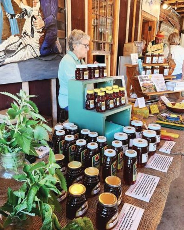 Honey jars on a burlap-covered market table in front of a farm market booth, with a vendor in a light blue shirt tending the display behind them.