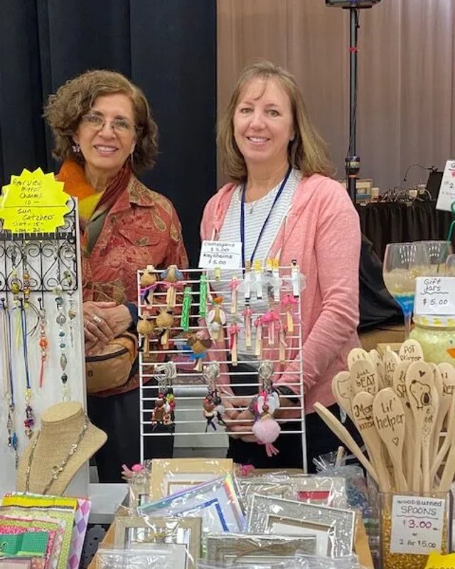 Two smiling women staffing a jewelry and crafts booth with necklaces and wooden utensils on display.