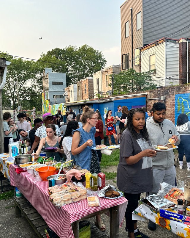 People gathering around a food table with plates and containers of food at the Brewerytown Garden, with murals on the walls behind.