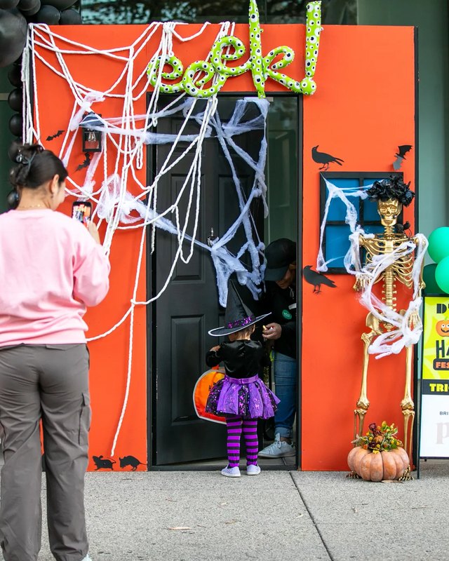 Costumed kid at a decorated Halloween trick-or-treat booth with a spider web door and a glowing 'peek' sign, with an adult partly visible at left.