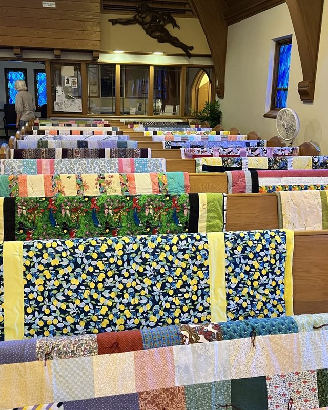 Handmade quilts draped over wooden pew backs in a church sanctuary, with stained glass windows and a wooden ceiling beyond.