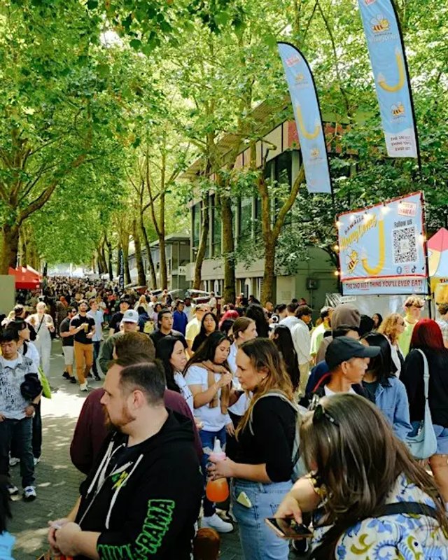 Large crowd of shoppers walking along a tree-lined street past market booths and banners at an outdoor market.