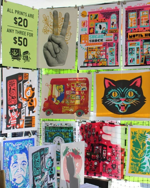 Colorful prints and posters pinned on a wall display at a printers fair vendor booth, with a green "All prints are $20" price sign.